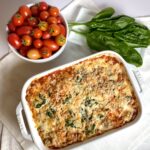 lasagna in white baking dish under fresh spinach, above right and fresh cherry tomatoes, above left