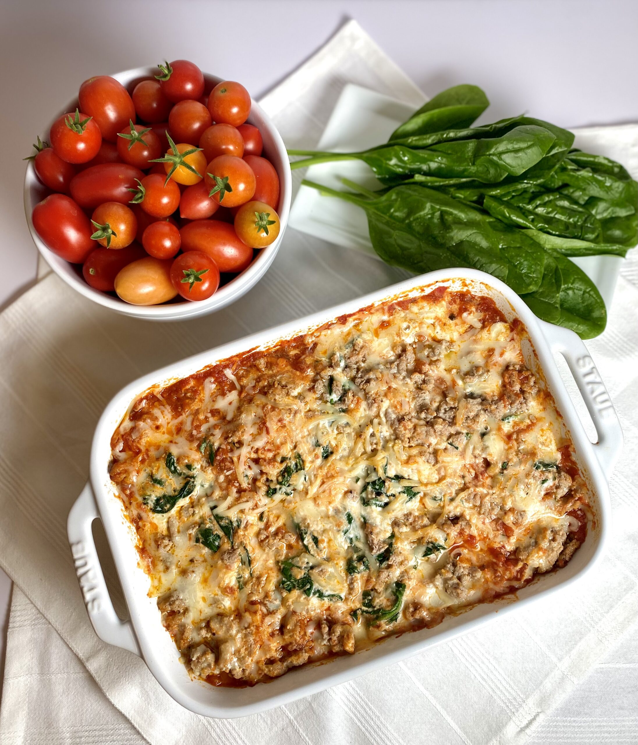 lasagna in white baking dish under fresh spinach, above right and fresh cherry tomatoes, above left