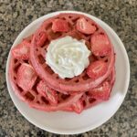 red velvet waffles stacked on white plate topped with dried strawberries and whipped cream