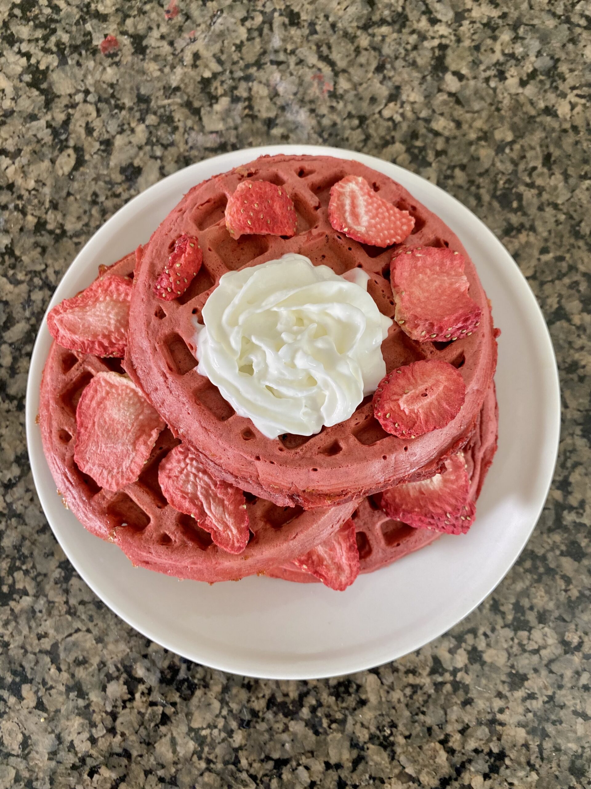 red velvet waffles stacked on white plate topped with dried strawberries and whipped cream