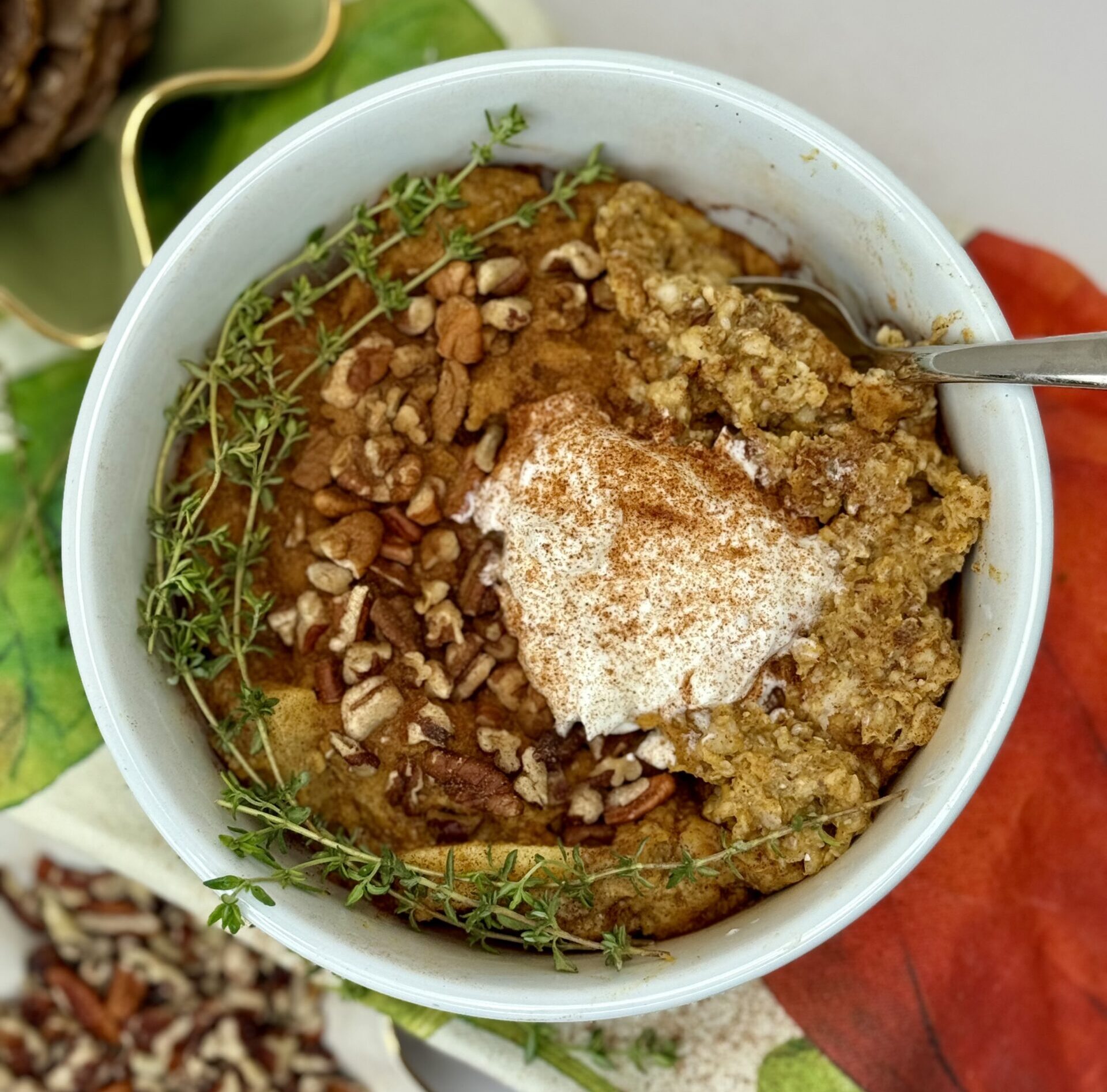 Pumpkin spice oatmeal in white bowl with garnish of Greek yogurt dollop and curved garnish of thyme, chopped pecans and cinnamon sprinkle
