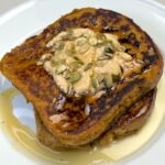 Two slices of Pumpkin Ricotta French Toast with pumpkin puree and greek yogurt cream topping and sprinkle of roasted pumpkin seeds and maple syrup