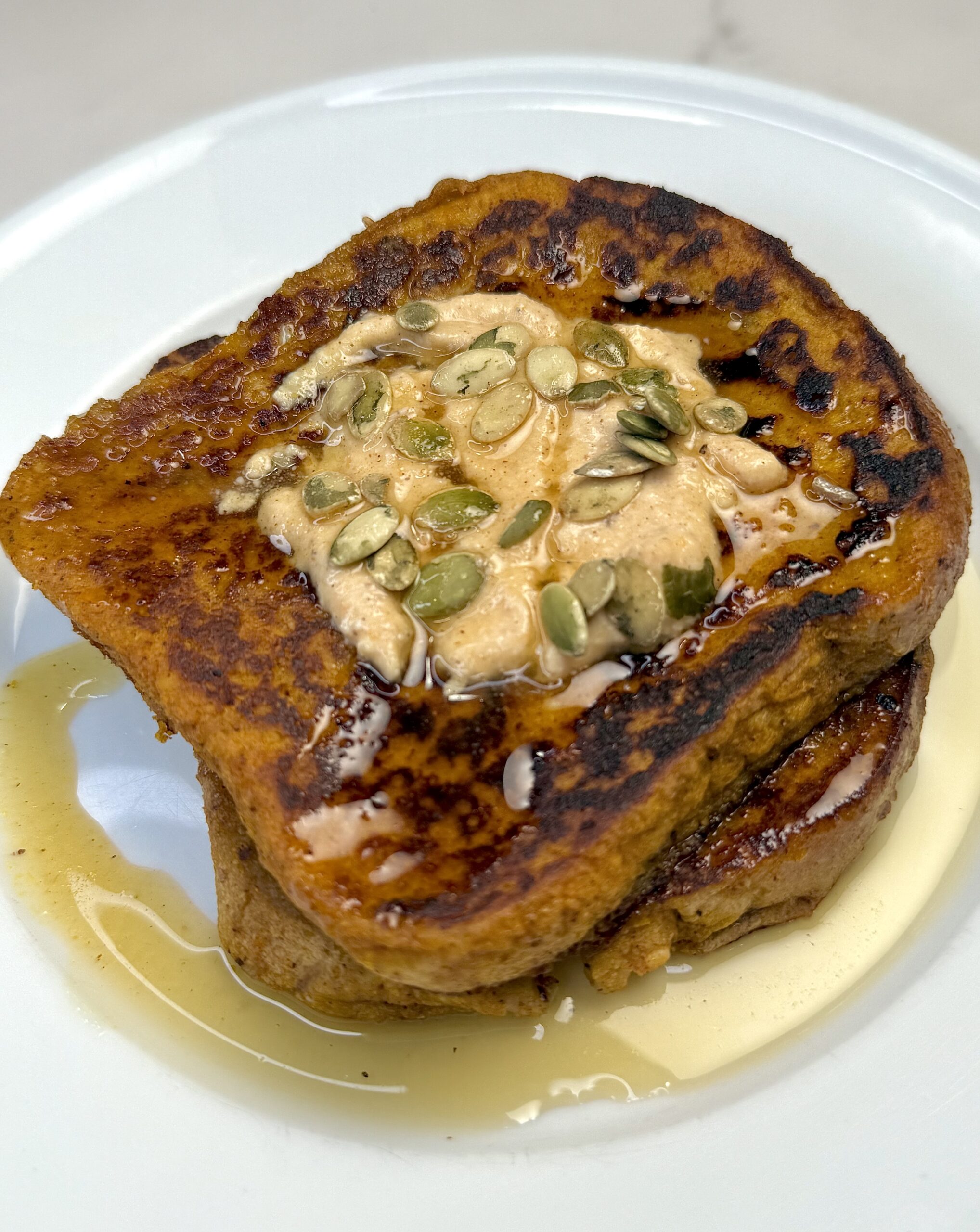 Two slices of Pumpkin Ricotta French Toast with pumpkin puree and greek yogurt cream topping and sprinkle of roasted pumpkin seeds and maple syrup