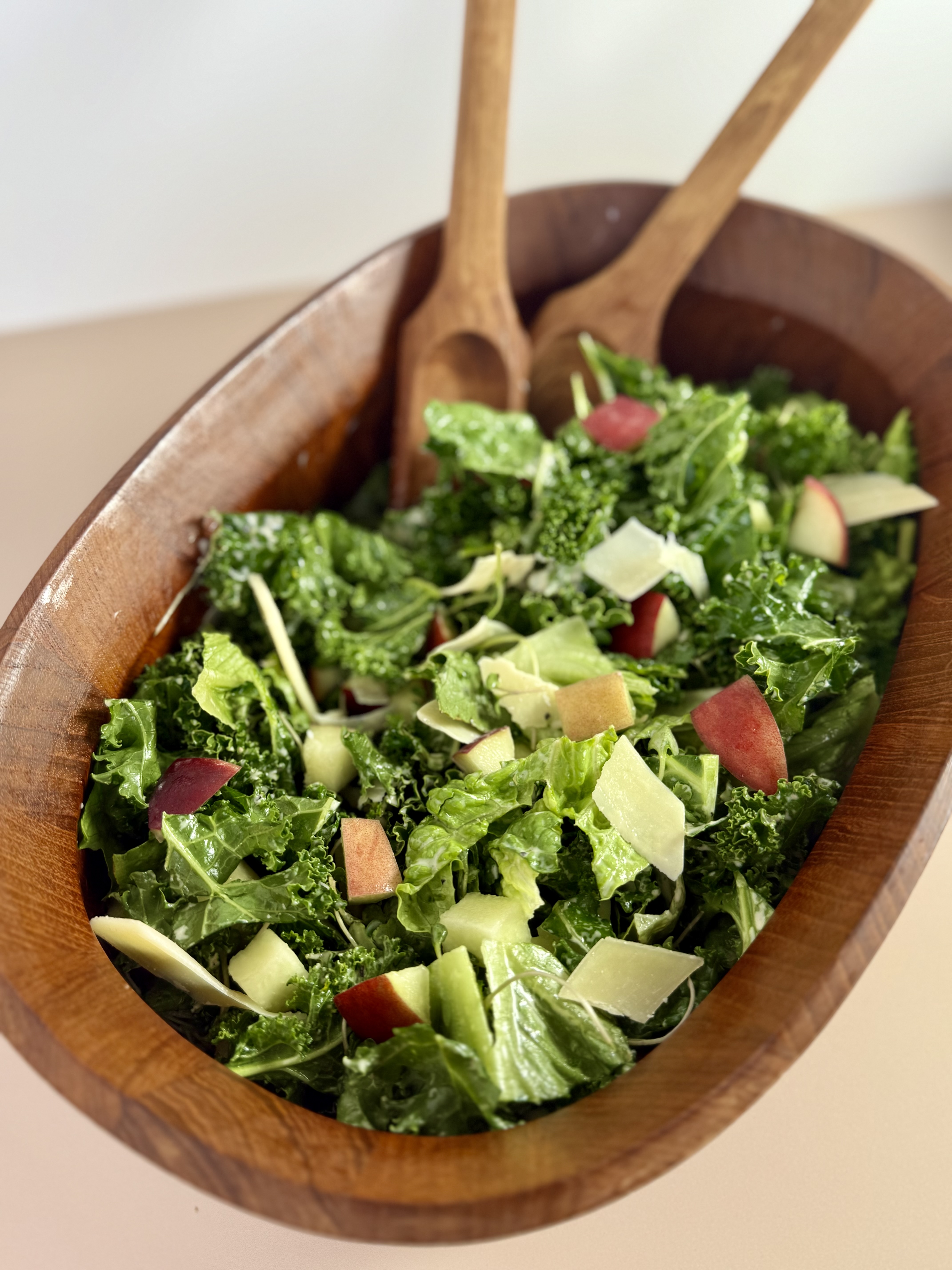white peach kale caesar and romaine lettuce salad in oval shaped wood bowl with 2 coordinating wood salad utensils