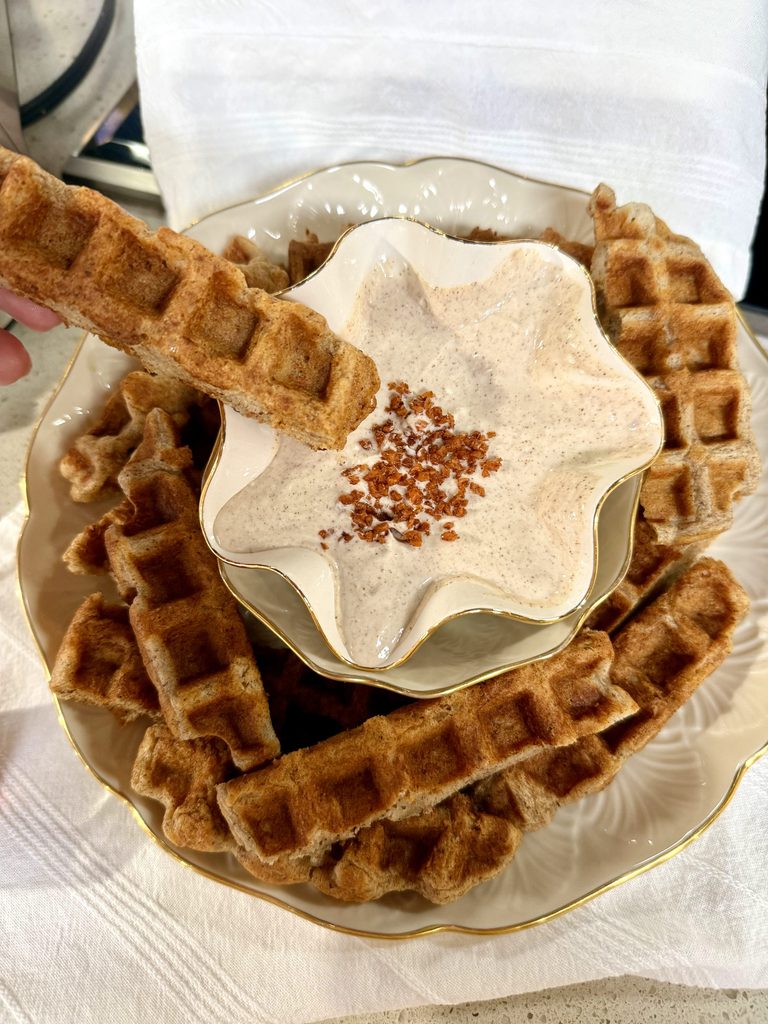 bowl of cinnamon, greek yogurt and cream cheese dip with carrot cake waffle sticks around it with one about to dip in
